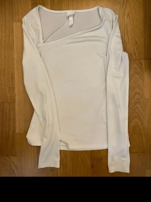 Vit topp från H&M - Säljer en stilren vit topp från H&M i stretchigt material. Toppen har lång ärm och en asymmetrisk halsringning som ger en modern vibe. Perfekt att matcha med jeans eller kjol för en clean look.