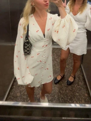 Vit blommig kortklänning från Adoore - Supersöt vit kortklänning från Adoore med små röda blommor, v-ringning och klädda knappar framtill. Klänningen har långa puffärmar och är gjord i ett strukturerat tyg som känns som bomull. Perfekt för sommarens alla tillfällen!