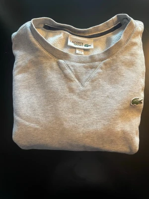Grå sweatshirt från Lacoste Sport - Snygg grå sweatshirt från Lacoste Sport i storlek S. Tröjan har klassisk rund hals och den ikoniska krokodilloggan broderad på bröstet. Perfekt för chill dagar och enkel att matcha med jeans eller joggers. Mjuk bomullskvalitet för extra komfort.
