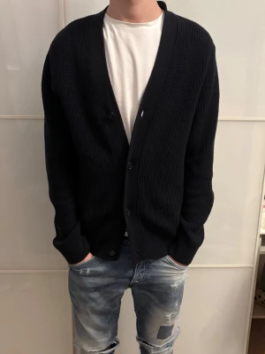 Svart ribbad kofta med knappar - Säljer en svart ribbad kofta med knappar framtill. Koftan har en djup v-ringning och långa ärmar. Perfekt att slänga över en t-shirt för en chill och stilren look. Passar både till jeans och andra byxor.