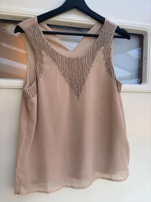 Beige blus med pärldetaljer - Säljer en beige blus från Only i lätt och luftigt material. Blusen är ärmlös och har snygga pärldetaljer framtill som bildar ett V-mönster. Perfekt för dig som gillar stilrena plagg med lite extra bling. Passformen är normal och toppen har en mjuk, feminin look.