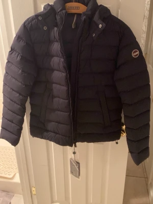 Mörkblå pufferjacka från Colmar - Säljer en mörkblå pufferjacka från Colmar Originals med huva och snygga horisontella quiltade paneler. Jackan har dragkedja framtill, två sidofickor och Colmar-logga på ärmen. Perfekt för kalla dagar och riktigt stilren look.