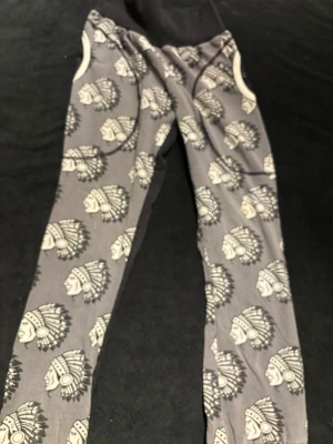 Mjukisbyxor Design Roshill, svart/grå, 140 - Unika mjukisbyxor från Design Roshill i storlek 140. Ena benet är helsvart och det andra är grått med coolt mönster av huvuden med fjäderskrud. Byxorna har resår i midjan och är gjorda i mjukt bomullstyg. Perfekta för chill dagar eller när du vill sticka ut lite extra.