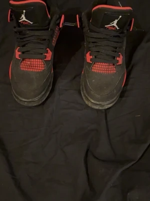 Nike Air Jordan 4 svart/röd sneakers - Säljer ett par Nike Air Jordan 4 sneakers i svart och rött med klassisk Jumpman-logga på plösen och sulan. Skorna har snörning, meshdetaljer och en platt sula. Perfekt för dig som gillar streetwear och vill sticka ut med en ikonisk modell.bra skick bara skitiga