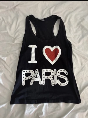 Svart linnetopp med I ♥ Paris-tryck - Svart linnetopp med stort vitt tryck 'I ♥ Paris' på framsidan. Hjärtat är rött och bokstäverna är dekorerade med silvriga nitar. 