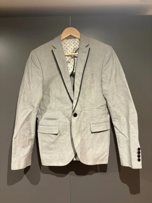 Ljusgrå kavaj från Selected Homme - Snygg ljusgrå kavaj från Selected Homme med svart kantband längs slaget och coolt mönstrat innerfoder. Klassisk passform med två fickor fram och knappar vid ärmslut. Perfekt för dig som vill ha en stilren och modern look. Storlek S