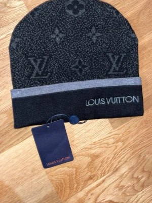 Mörkgrå  mössa med lyx design - Snygg mörkgrå mössa med klassiskt monogram och diskret logga broderad framtill. Mössan har en bred uppvikt kant i grått och är stickad i mjukt material som känns lyxigt och bekvämt.