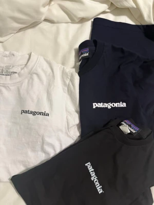 Patagonia t-shirt i svart, vit och marinblå - Säljer tre snygga Patagonia t-shirts i svart, vit och marinblå. Alla har klassisk logga tryckt på bröstet, och en av de svarta har även ett stort färgglatt logotryck på ryggen. T-shirtsen är kortärmade och gjorda i mjuk bomull med normal passform.