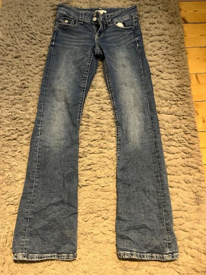 Blå bootcut jeans med klassisk tvätt - Snygga blå jeans med bootcut-snitt och klassisk tvätt. Modellen har fem fickor, bälteshällor och knappgylf. Jeansen är tillverkade i slitstarkt denim och har en lätt urtvättad look framtill och baktill. Perfekta för dig som gillar retrostil och vill ha ett par jeans med lite extra vidd vid benslutet.