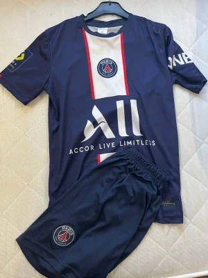 PSG Neymar 10 fotbollströja och shorts - Säljer ett ORIGINAL mörkblått PSG matchställ med Neymar 10-tryck på ryggen. Setet består av en kortärmad tröja med vita och röda detaljer samt klubbmärke på bröstet och ett par matchande shorts med PSG-logga. Perfekt för dig som älskar fotboll och vill ha en snygg look på planen.