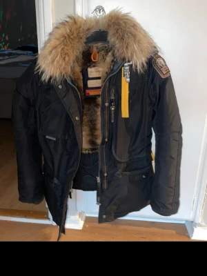 Svart Parajumpers parkas med päls - Säljer en svart Parajumpers parkas med stor fluffig päls runt huvan och mjukt foder. Jackan har flera fickor, gul detalj vid dragkedjan och patch på ärmen. Perfekt för kalla dagar och riktigt snygg streetstil. Hål och fläckfri och väldigt sparsamt använd 