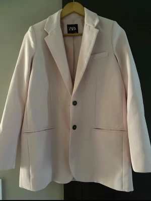 Ljusrosa kavaj från Zara - Snygg ljusrosa kavaj från Zara med klassisk krage och två svarta knappar framtill. Kavajen har tre fickor och är tillverkad i ett mjukt material som ger en stilren look. Perfekt för dig som vill ha en fräsch och modern vibe.