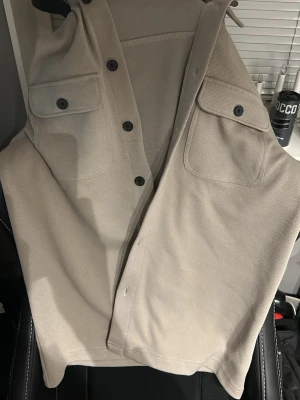 Beige väst med fickor och knappar - Säljer en stilren beige väst med två bröstfickor med lock och svarta knappar framtill. Västen har en rak passform och är tillverkad i ett kraftigare bomullstyg som ger en clean och modern look. Perfekt att slänga över en hoodie eller t-shirt.