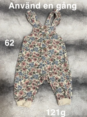 Blommig byxdress med knappar, stl 62 - Supersöt byxdress med blommigt mönster i rosa, blått och grönt på ljus botten. Byxdressen har knappar framtill och axelband med volangdetalj. Muddar vid benslut och tryckknappar för enkel påklädning. Perfekt för små barn som vill vara både bekväma och stiliga.🌸 Använda en gång. Storlek 62.