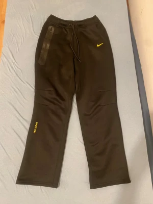 Svarta NOCTA Nike mjukisbyxor - Svarta mjukisbyxor från Nike NOCTA med gul logga på benet och gul swoosh på höften. Byxorna har snörning i midjan, en lång dragkedja på ena fickan och är tillverkade i mjukt bomullsmaterial. Snygg loose passform och stilren design.