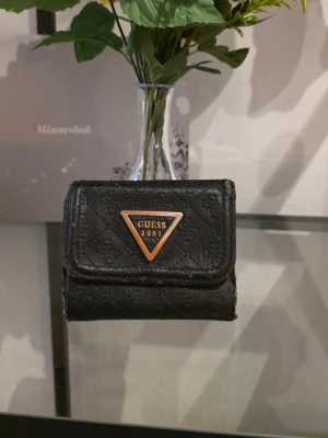 Svart plånbok från Guess - Snygg svart plånbok från Guess med klassiskt triangelformat metallogo framtill. Plånboken har ett diskret mönster med Guess-loggor över hela ytan och är tillverkad i syntetmaterial. Perfekt storlek för kort och kontanter, smidig att ha med sig överallt.