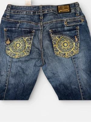 Blå jeans från Baby Phat med broderi - Säljer mina älskade blå jeans från Baby Phat med guldiga broderade detaljer på bakfickorna och dekorativa knappar fram. Jeansen har låg midja och klassisk bootcut-modell. Materialet är slitstarkt jeans med snygg tvätt och guldfärgade detaljer som sticker ut.