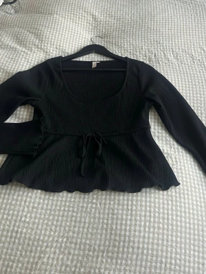 Svart topp med knyt - Säljer en svart långärmad topp med  knyt i midjan. Toppen har en rundad urringning och lätt utsvängd nederkant som ger en söt siluett. Perfekt att styla med jeans eller kjol för en trendig look.
