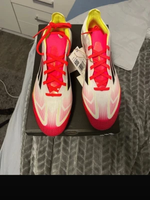 Adidas F50 PRO FG - Säljer ett par Adidas f50 PRO fotbollsskor med snygg design i vitt, rött och gult. Skorna har svarta ränder på sidan, röda snören och en platt sula med dobbar för gräsplan. Ovandelen är i syntetmaterial och insidan har neonfärgade detaljer. Perfekt för dig som vill sticka ut på planen.