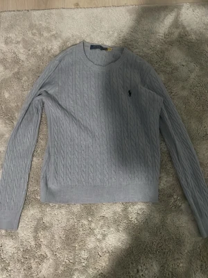 Ljusgrå kabelstickad tröja Polo Ralph Lauren - Säljer en ljusgrå kabelstickad tröja från Polo Ralph Lauren. Tröjan har rund hals, långärmade och ett broderat mörkblått logomärke på bröstet. Materialet är mjuk bomull och passformen är normal. Perfekt för dig som gillar klassisk och stilren look. Storlek XS, hör av er vid frågor. Aldrig använd bata testad. Den har inte stickar material det är ett lent material