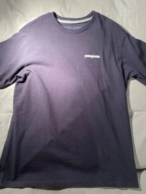 Mörkblå Patagonia t-shirt med tryck - Säljer en mörkblå t-shirt från Patagonia nyskick i regular fit storlek S. Framsidan har en liten vit Patagonia-logga och baksidan har ett stort färgglatt tryck med berg och logotyp. T-shirten är gjord av 50% återvunnen polyester och 50% återvunnen bomull. Perfekt för dig som gillar outdoor-vibbar.