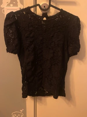 Svart spetsblus med kort ärm - Säljer en svart blus i spets med blommigt mönster och korta ärmar. Storlek m men passar s Toppen har en liten knapp i nacken och är lätt transparent tack vare spetsmaterialet. Perfekt för dig som gillar detaljer och en romantisk vibe. Har klippt ärmarna själv så lite ojämnt och trådig men syns inte så mycket