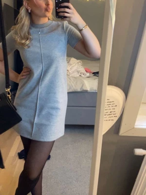 Grå stickad kortklänning från Zara - Superfin grå stickad kortklänning från Zara med rund halsringning och korta ärmar. Klänningen har en rak passform och en diskret söm framtill som ger en clean look. Perfekt att styla med strumpbyxor och boots för en snygg vardagsstil.