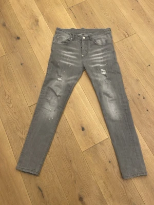 Grå slim fit jeans från Dsquared2 - Säljer ett par grå slim fit jeans från Dsquared2 med slitningar och fina detaljer som färgstänk och distressed look. Jeansen har klassisk femficksdesign, knappgylf och Dsquared2-logga på bakfickan. Perfekt för någon som festar och vill sticka ut.