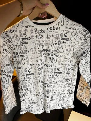 Vit långärmad topp Pantaloons Junior - Cool långärmad topp från Pantaloons Junior i vitt med svart textprint över hela plagget. Trycket består av olika ord och uttryck i handskriven stil som ger en streetig vibe. Tillverkad i mjuk bomull och har en klassisk rund halsringning.