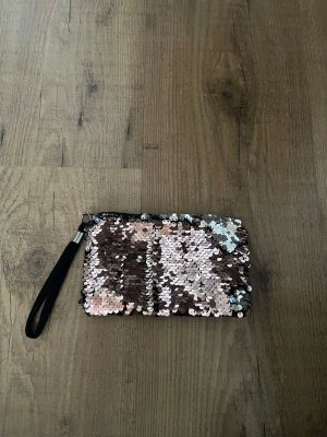 Glittrig väska med paljetter - Säljer en glittrig liten väska med paljetter i silver och rosa toner. Väskan har en svart handledsrem och dragkedja upptill. Paljetterna kan vändas för att ändra färg mellan silver och rosa. Väskan är i väldigt bra skick, väskan är 17 cm lång och 10 cm bred.