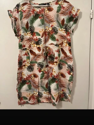 Blommig kortklänning med snörning - Säljer en färgglad kortklänning med tropiskt blommönster i grönt, rött, beige och svart. Klänningen har snörning i midjan och korta ärmar, perfekt för varma dagar. Materialet känns lätt och luftigt, och den har en avslappnad passform.