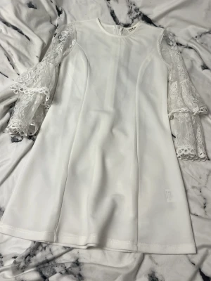 Vit klänning med spetsärmar  - Supersnygg vit kortklänning från Shein med unika spetsärmar i två lager och rund hals. Klänningen har en stilren, rak passform och dragkedja i ryggen. Materialet är mjukt och slätt, medan ärmarna är gjorda av transparent spets med blommönster och dekorativa kanter.