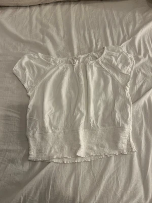 Vit blus från KappAhl - Säljer en vit blus från KappAhl i storlek 158/164. Blusen har korta ärmar, rund halsringning med liten knyt-detalj och ett brett smockparti nedtill. Materialet är lätt och krinklat, perfekt för varma dagar.