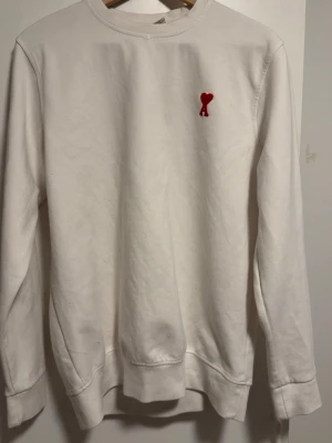 Vit sweatshirt från Ami Paris - Säljer en vit sweatshirt från Ami Paris med rött broderat hjärta och A på bröstet. Tröjan har rund hals, långa ärmar och ribbade muddar vid ärmslut och nederkant. Perfekt för dig som gillar stilrena och enkla plagg med en liten detalj.