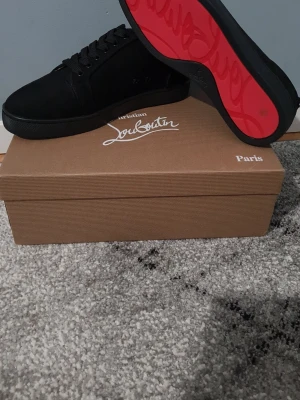 Svarta sneakers från Christian Louboutin - Säljer ett par svarta sneakers från Christian Louboutin med klassisk röd sula och snygg snörning. Skorna har rund tå och är tillverkade i mocka med detaljer i skinn. Kommer med originalkartong och dustbag med logga.