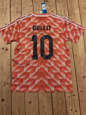 Holländsk retro fotbollströja Adidas - Säljer en ikonisk orange retro fotbollströja från Adidas med KNVB-logga och svart Adidas-märke på bröstet. Tröjan har ett geometriskt mönster, vita ränder på axlarna och V-ringad krage. På ryggen står det 'Gullit' och nummer 10 i svart.