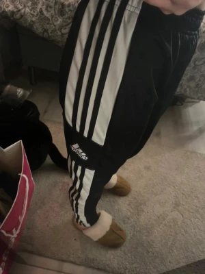 Svarta Adidas byxor med vita ränder – med defekter - Svarta Adidas byxor med klassiska vita ränder längs benen och Adidas-logga framtill. Bekväm modell med resår i midjan. Observera att byxorna har defekter: de har fått svart färg på sig som inte går bort i tvätten. Detta syns på vissa partier. Passar bra som vardags-, hemma- eller träningsbyxor där utseende inte är det viktigaste.