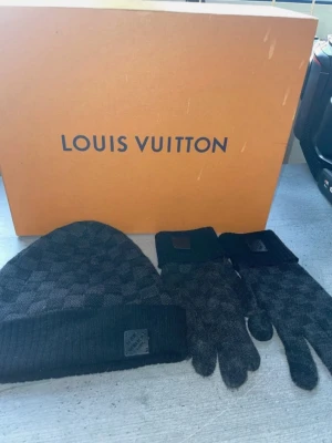 Louis Vuitton rutig mössa & vantar - Säljer ett set från Louis Vuitton med mössa och vantar i mörkgrått och svart rutigt mönster. Båda har ribbade muddar och en metallbricka med Louis Vuitton-logga. Materialet är mjuk ull och kashmir, perfekt för kalla dagar.