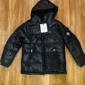 Svart pufferjacka från Moncler S - Säljer en svart pufferjacka från Moncler i storlek S. Jackan har huva, två snedställda fickor med dragkedja och Moncler-logga på ärmen. Fylld med vitt gåsdun för extra värme och har en glansig finish. Perfekt för kalla dagar och riktigt snygg streetstil.