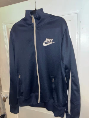 Marinblå Nike zip hoodie - Säljer en marinblå zip hoodie från Nike med vit dragkedja och logga på bröstet. Tröjan har högt krage, långa ärmar med vita ränder längs axlarna och två fickor med dragkedja. Perfekt för dig som gillar sportig och clean stil.