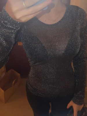 Glittrig svart långärmad topp - Säljer en svart långärmad topp med glittrigt material och rund halsringning. Toppen är tight och har en snygg, figurnära passform som verkligen framhäver siluetten. Perfekt för att sticka ut med lite extra shine. Toppen är igenom synlig och ger lite ev en intimissimi vibe. Tunnt material och perfekt ifall man ska klä upp sig 