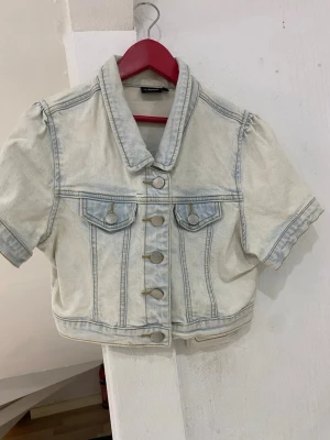 Ljus jeansjacka med korta ärmar - Snygg ljusblå jeansjacka från Very Bonita med korta ärmar och croppad passform. Jackan har klassisk krage, silverfärgade knappar framtill och två bröstfickor med lock. Perfekt för dig som vill ha en chill och trendig look.