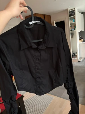 Svart croppad skjorta med knappar - Säljer en svart croppad skjorta från NA-KD med långa ärmar och knappar framtill. Skjortan har klassisk krage och coola rynkade detaljer längs ärmarna. Perfekt till jeans eller kjol för en trendig look.