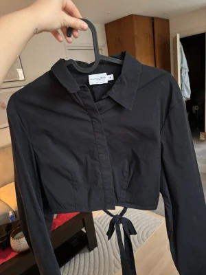Svart croppad skjortblus med knyt - Säljer en svart croppad skjortblus från NAKD i storlek 36. Toppen har klassisk krage, långa ärmar och knytband i midjan för en snygg öppen rygg detalj. Tillverkad i bomull och har en stilren look som passar till mycket.