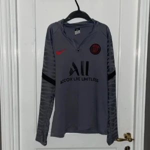 Grå långärmad PSG fotbollströja Nike - Snygg grå långärmad fotbollströja från PSG med svarta och röda detaljer. Tröjan har halv dragkedja, diskret mönster på ärmarna och klubbmärke på bröstet. Perfekt för dig som älskar fotboll och vill sticka ut på planen.