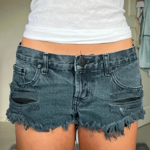 Svarta jeansshorts med slitningar från One tabelspoon - Snygga svarta jeansshorts från One Teaspoon med låg midja, slitna detaljer och fransig kant. Klassisk femficksmodell med knapp och dragkedja framtill. Perfekta för en edgy och avslappnad stil.