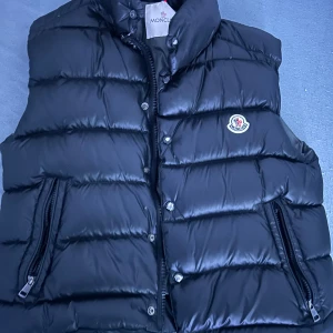 Svart dunväst från Moncler - Säljer en svart dunväst från Moncler med quiltad design och hög krage. Västen har två praktiska fickor med dragkedja och klassisk Moncler-logga på bröstet. Insidan har en unik serietidningsetikett och märkning. Perfekt för att hålla värmen med stil.