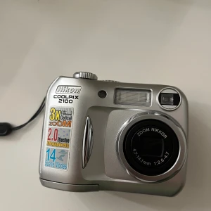 Coolpix 2100 - Nikon Coolpix 2100 digitalkamera med 3x optisk zoom, 2.0 megapixlar och 14 scenlägen. Kameran är i fint skick med minimalt slitage, batterifack och CF-kortplats synliga. Perfekt för dig som vill ha en kompakt och pålitlig kamera. OBS! CF-kortplats saknas samt laddare. Gammal kamera vet ej om det funkar. 