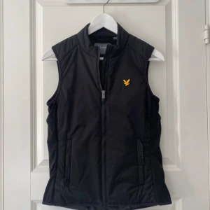 Svart dunväst från Lyle & Scott S - Svart dunväst från Lyle & Scott med gul broderad logga på bröstet. Västen har dragkedja framtill och två fickor med dragkedja. Snygg, enkel design som funkar till många outfits. Materialet är syntet och västen har en klassisk passform.