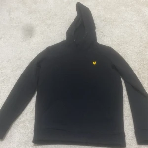 Svart hoodie från Lyle & Scott - Svart hoodie från Lyle & Scott med klassisk gul logga på bröstet. Tröjan har huva, långa ärmar och en stor magficka framtill. Perfekt för dig som gillar en enkel och stilren look.pris kan diskuteras privat 